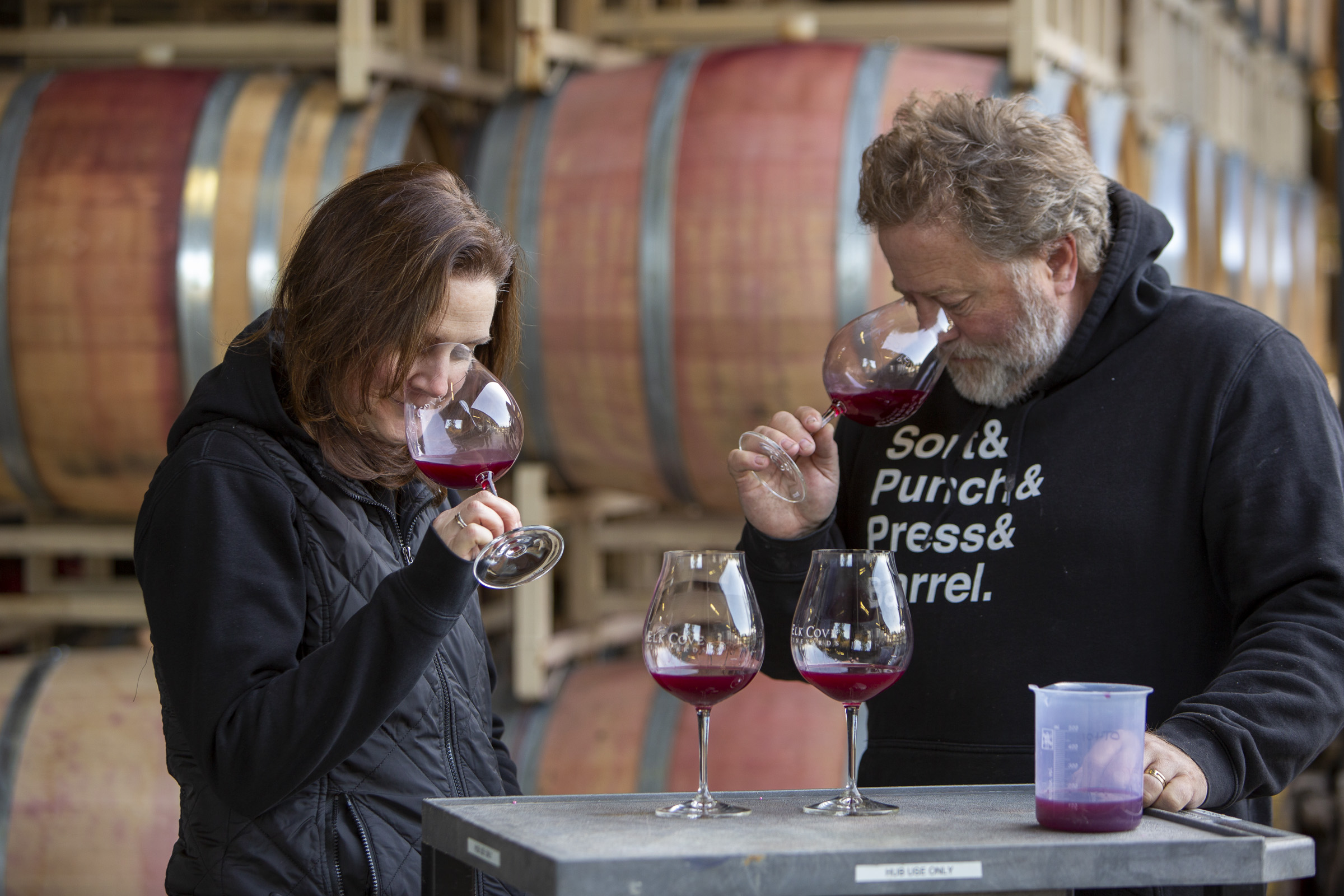 two winemakers, Adam Campbell and Heather Perkin smelling red wine samples in front of barrels